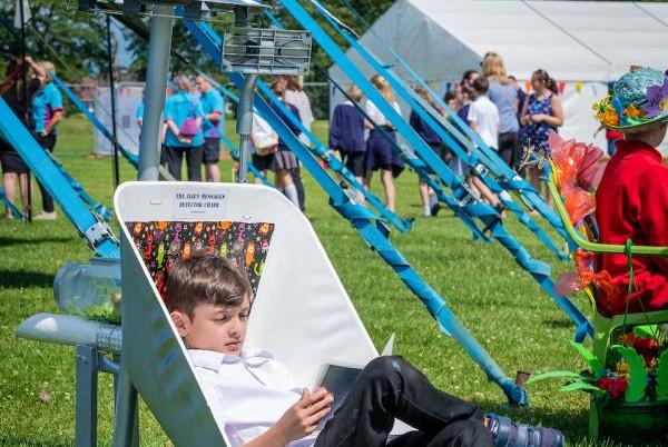 boy sat in a chair at the festival. He's reading, there's a crowd of people in the background.