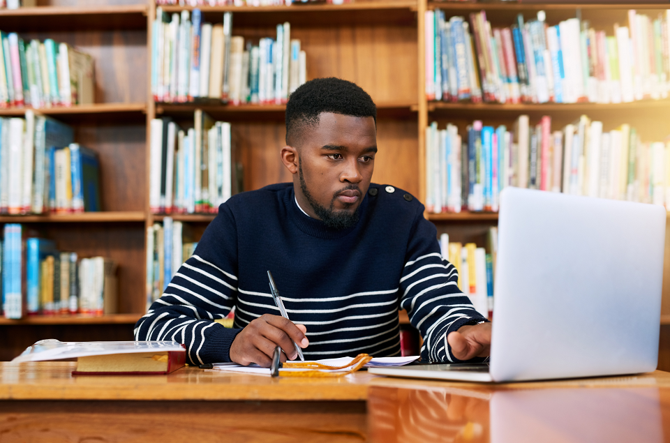 man working at a laptop in a reference library. behind him are shelves with books on them.